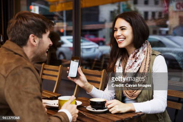 happy woman showing mobile phone to boyfriend while enjoying coffee at cafe - dating show stock pictures, royalty-free photos & images