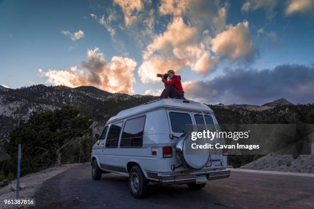 woman photographing while sitting on car roof against sky during sunset - nevada stock-fotos und bilder