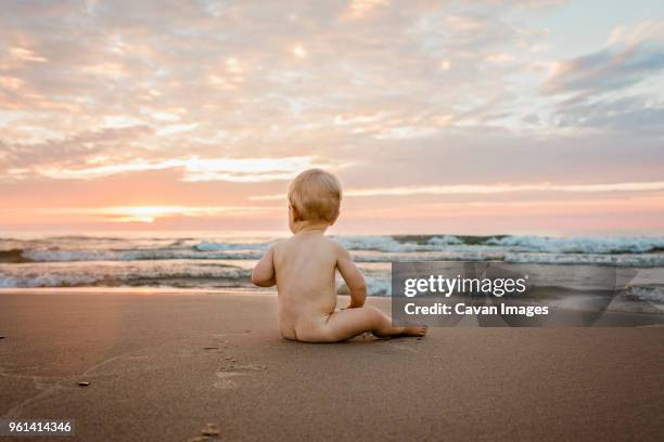 full length of naked baby boy sitting on sand at beach against cloudy sky during sunset - boys bare bum stock pictures, royalty-free photos & images