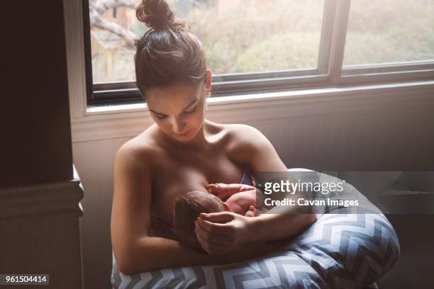 mother breastfeeding son while sitting by window at home - sucking fotografías e imágenes de stock