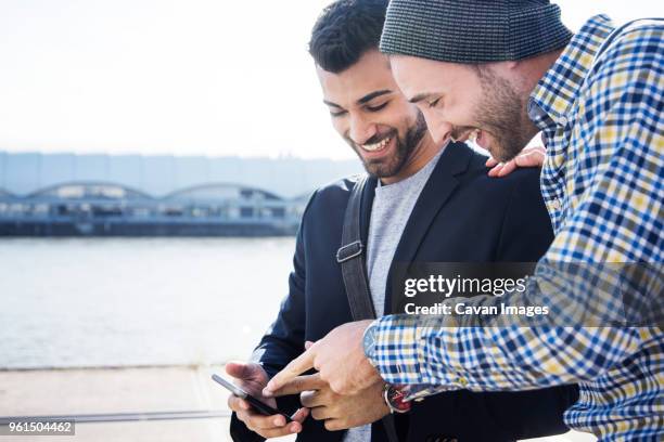 happy friends using phone while standing by canal against sky - cell mates stock-fotos und bilder