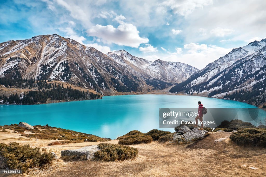 Full length of backpacker looking at view while standing by lake against mountains and cloudy sky during winter