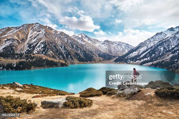 full length of backpacker looking at view while standing by lake against mountains and cloudy sky during winter - kazakistan foto e immagini stock