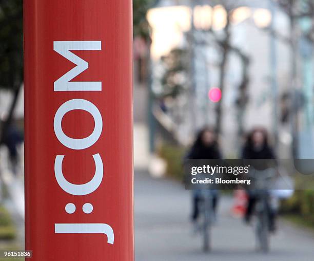 Women cycle behind a sign for Jupiter Telecommunications Co.'s J:COM shop in Tokyo, Japan, on Monday, Jan. 25, 2010. KDDI Corp., Japan's...