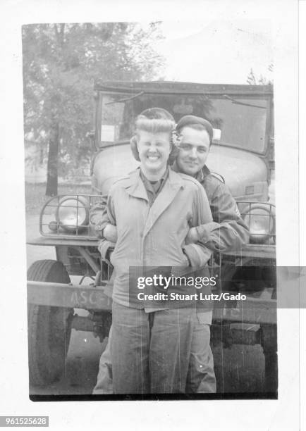 Black and white photograph, showing a man behind a woman, both in three-quarter length view, wearing military uniforms, and standing in front of a...