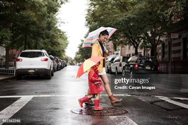 side view of mother and daughter holding umbrellas while crossing road - girl smiling on rain stock-fotos und bilder