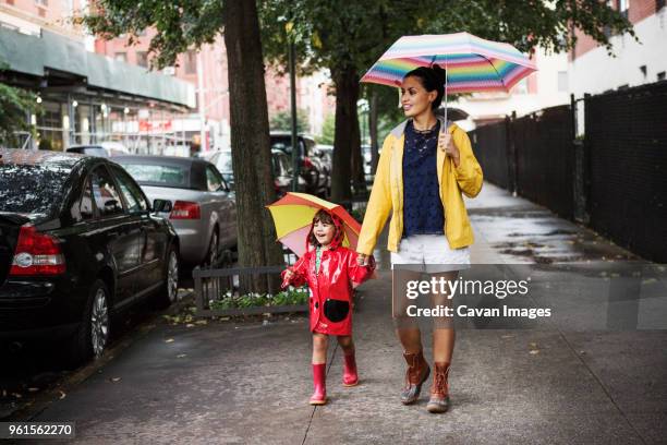 happy mother and daughter holding umbrellas while walking on sidewalk - mother protecting from rain stock-fotos und bilder