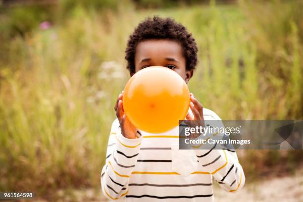 boy blowing balloon while standing against grass field - inflar fotografías e imágenes de stock