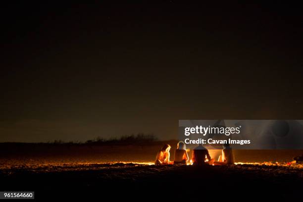 friends sitting around bonfire at beach during night - holzfeuer stock-fotos und bilder