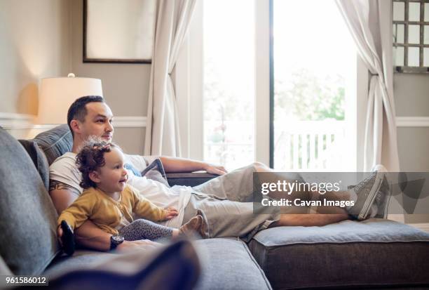 father and daughter watching television while sitting on sofa at home - genderblend2015 stock pictures, royalty-free photos & images