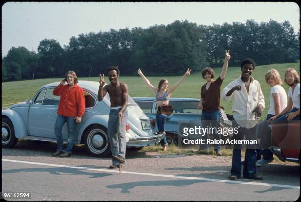 View of a group of people, by several parked cars on the side of the road, on their way to attend the Woodstock Music and Arts Fair, Bethel, New...