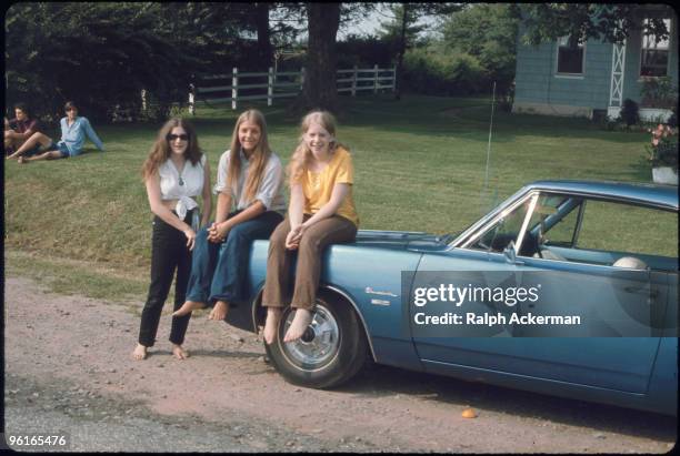 Portrait of three unidentiied and barefoot women, two of whom are seated on the hood of a Plymouth Barracuda parked on the side of a gravel road near...