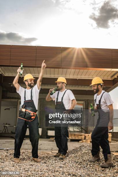 full length of happy manual workers drinking beer at construction site. - beer helmet stock pictures, royalty-free photos & images