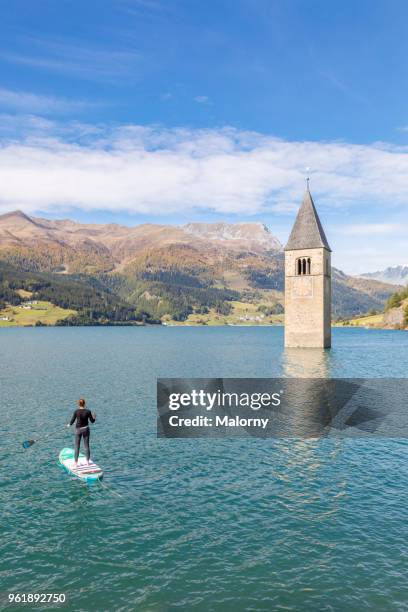 young woman paddleboarding near steeple and bell tower of a church sunken in lake reschen, reschensee or lago di resia, south tyrol, italy - reschenpass stock-fotos und bilder