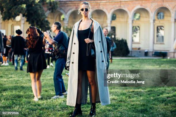 Model Hailey Clauson wears black round sunglasses, a tan trench coat on her shoulders, and black sleeveless dress, purse, and boots during Milan...