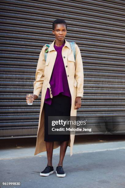 Model Achok Majak holds a glass of water and wears a long tan jacket, purple top, black skirt, and black slip on shoes during Milan Fashion Week...