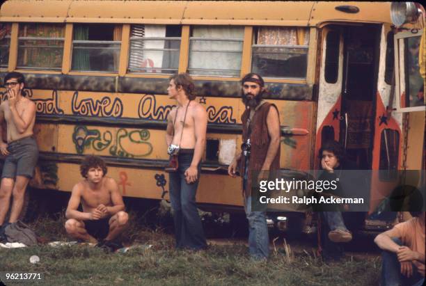 Near the 'Free Stage' at the Woodstock Music and Arts Fair, several men, two with cameras around their necks, lean against a decorated school bus...