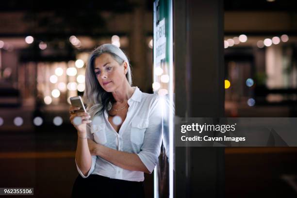businesswoman using phone while leaning on bus stop poster in city - night before fotografías e imágenes de stock