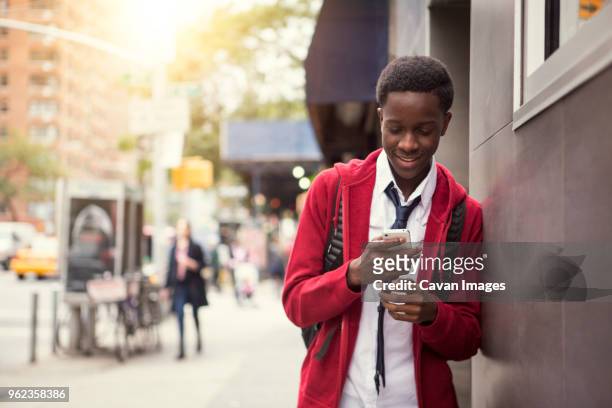 smiling student using phone while leaning on wall in city - school tie stock-fotos und bilder