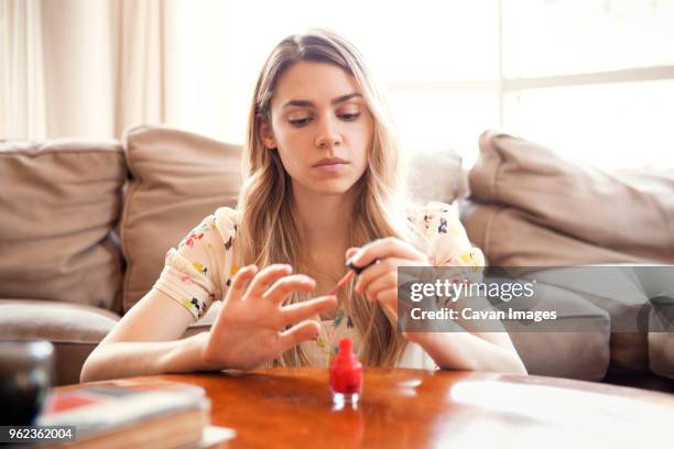 woman applying nail polish while sitting against sofa at home - fingernägel lackieren stock-fotos und bilder