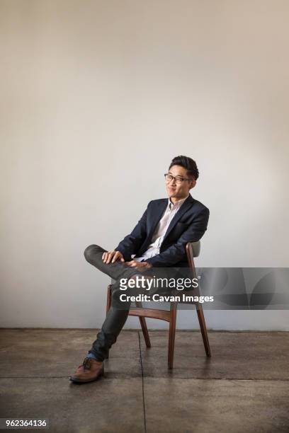 full length of smiling confident businessman sitting on chair against wall in office - sedia foto e immagini stock
