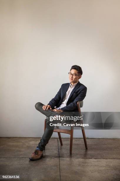 full length of smiling confident businessman sitting on chair against wall in office - asian man sitting at desk stock pictures, royalty-free photos & images