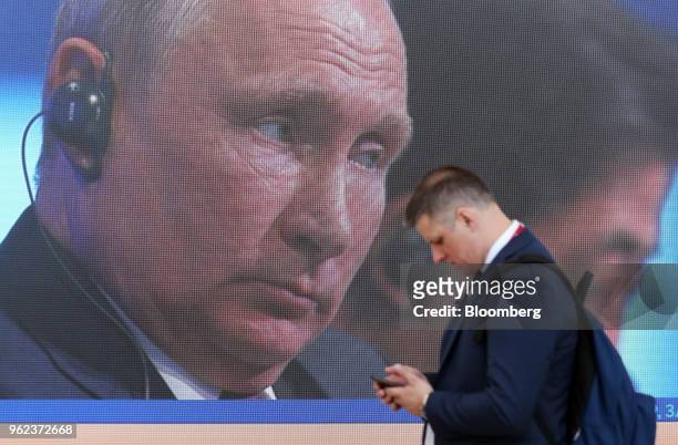 An attendee checks his smartphone as he walks past a screen showing the plenary session speech by Vladimir Putin, Russia's president, at the St....