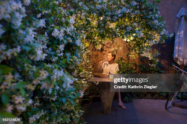 woman sitting under star jasmine pergola at dusk, enjoying her book and glass of wine - italian woman stock-fotos und bilder