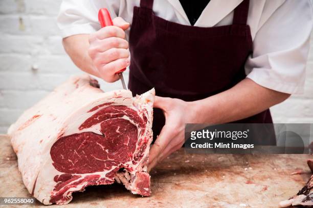 close up of butcher wearing apron standing at a wooden butchers block, butchering beef forerib. - butcher foto e immagini stock