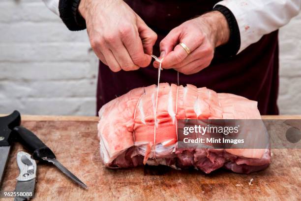 close up of butcher wearing apron standing at a wooden butchers block, preparing a rolled pork belly using string. - wooden block stockfoto's en -beelden