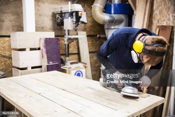 man wearing ear protectors, protective goggles and dust mask standing in a warehouse, using sander to smoothen piece of wood. - wood ear stock pictures, royalty-free photos & images