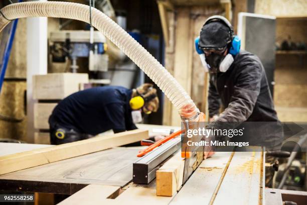 man wearing ear protectors, protective goggles and dust mask standing in a warehouse, cutting piece of wood with circular saw. - wood ear stock pictures, royalty-free photos & images