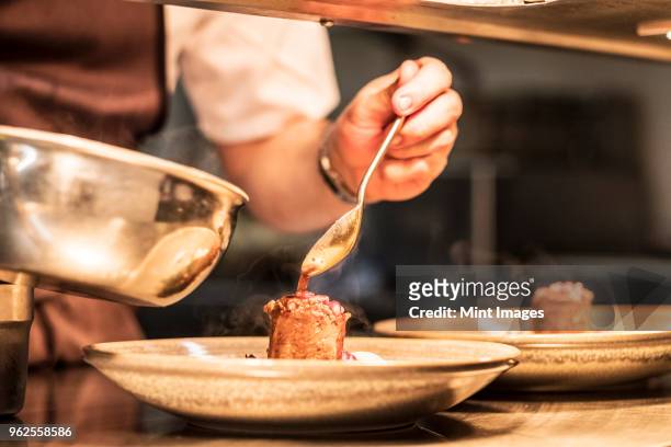 close up of chef in commercial kitchen holding spoon, drizzling sauce on plate. - spoon in hand stock pictures, royalty-free photos & images