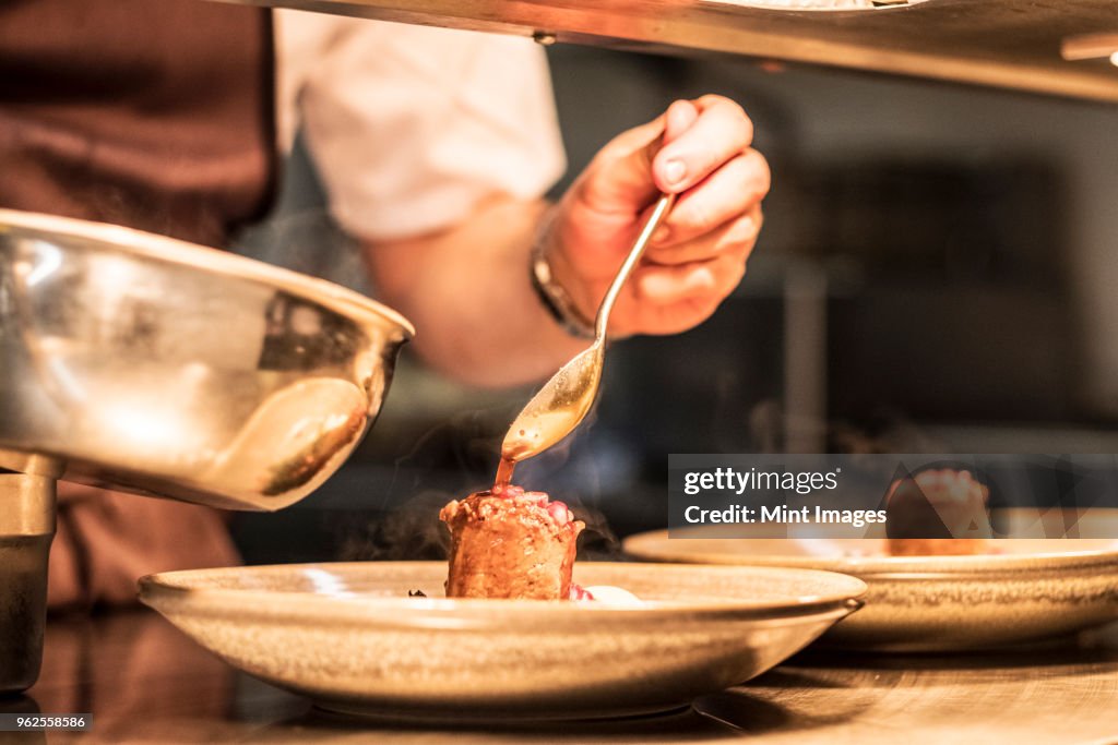 Close up of chef in commercial kitchen holding spoon, drizzling sauce on plate.