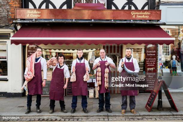 group of men, butchers and fishmonger, wearing aprons, standing on the pavement outside a butcher shop, holding pieces of meat and fish, looking at camera. - metzger stock-fotos und bilder