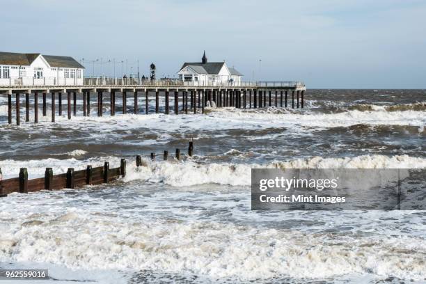seascape with waves rolling onto beach near groyne and white wooden buildings on a pier. - southwold stockfoto's en -beelden