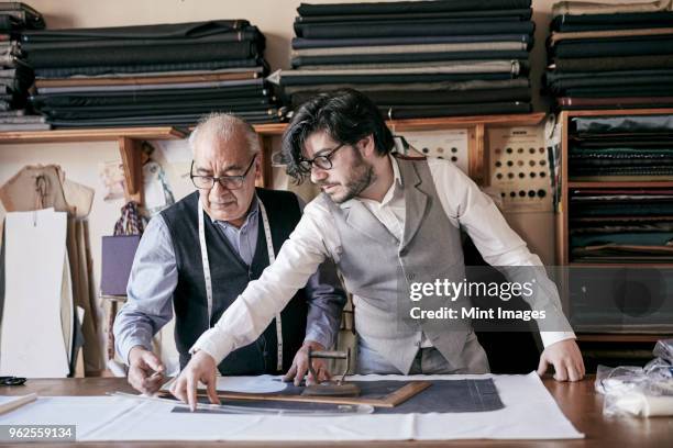 a tailor and a younger man, an assistant or apprentice working together, measuring cloth and cutting patterns. - family business generations imagens e fotografias de stock