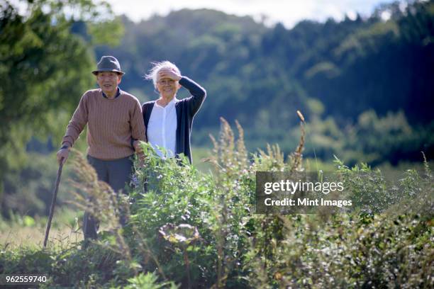 husband and wife, elderly man wearing hat and using walking stick and elderly woman walking along path. - japanese senior couple stockfoto's en -beelden