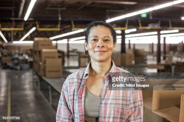 portrait of an african american female warehouse worker in a large distribution warehouse with products stored in cardboard boxes. - blue collar portrait stock pictures, royalty-free photos & images