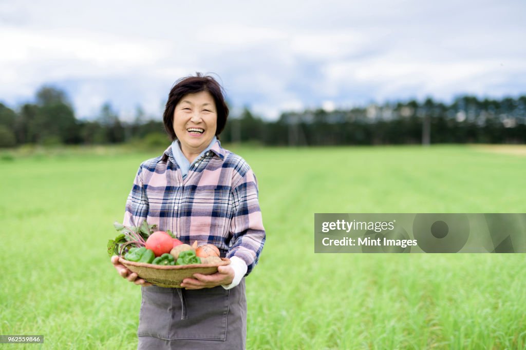 Woman with black hair wearing checkered shirt standing in a field, holding basket with fresh vegetables, smiling at camera.