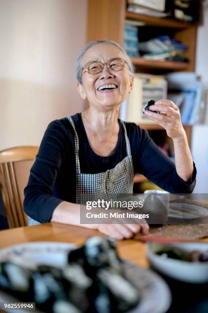 elderly woman sitting at kitchen table, eating sushi, smiling at camera. - japanese old man stock-fotos und bilder