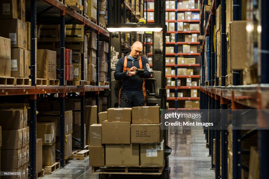 Warehouse worker wearing a safety harness while operating a motorized stock picker in an aisle between large racks of cardboard boxes holding product on pallets in a large distribution warehouse.