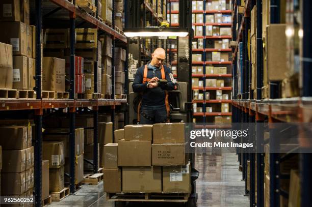 warehouse worker wearing a safety harness while operating a motorized stock picker in an aisle between large racks of cardboard boxes holding product on pallets in a large distribution warehouse. - safety equipment stock pictures, royalty-free photos & images