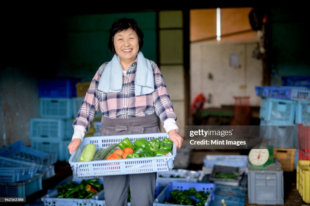 Woman with black hair wearing checkered shirt standing in front of barn, holding blue plastic crate with fresh vegetables, smiling at camera.