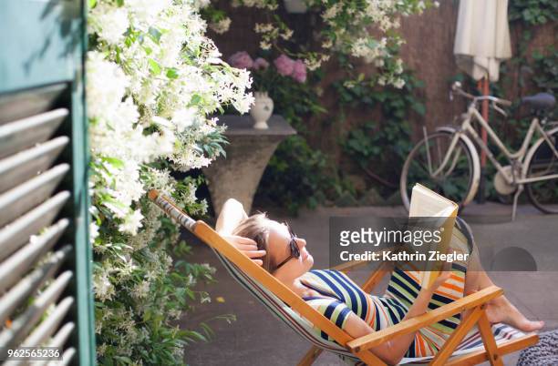 woman relaxing on deck chair in backyard, reading a book - reading outside stock pictures, royalty-free photos & images