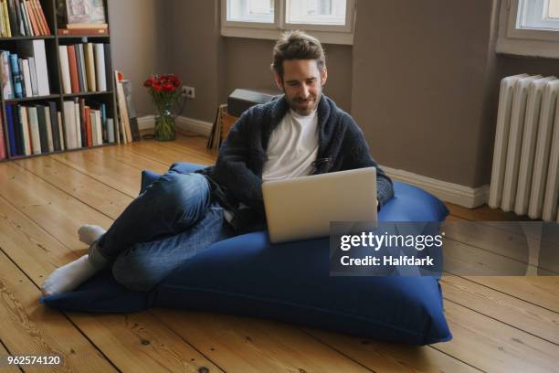 full length of man sitting with laptop on blue bean bag in room at home - bean bags fotografías e imágenes de stock