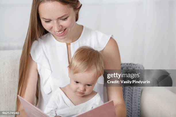 smiling mother and daughter reading picture book sitting on sofa at home - pindyurin foto e immagini stock