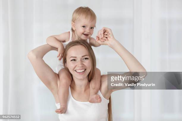 happy mother carrying daughter on shoulders against white curtain at home - pindyurin stock-fotos und bilder
