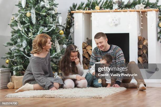 happy family sitting on rug by christmas tree at home - pindyurin stock-fotos und bilder