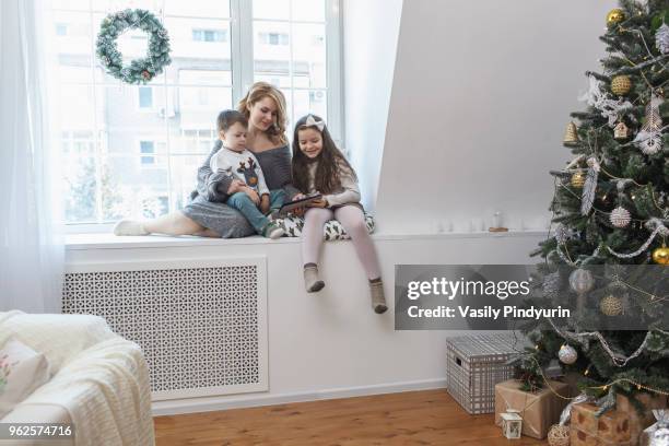 woman sitting with children on window sill at home during christmas - pindyurin foto e immagini stock